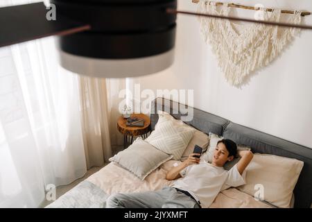 Asian boy smiling and using mobile phone while resting on bed at home Stock Photo