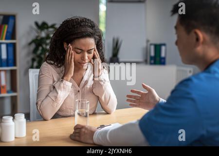 Serious adult asian male doctor calms sad young african american female patient in clinic office interior Stock Photo