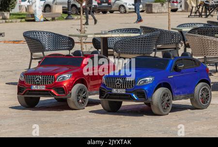 Budvar, Montenegro- April 26th, 2022 -Two miniature Mercedes children's toys parked in the harbour by some garden chairs Stock Photo