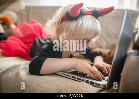 A little girl in a costume with Halloween horns writes a message on a laptop. Chat online for the holidays. Life style Stock Photo