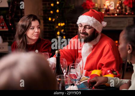 Husband wearing santa claus costume celebrating christmas with wife and parents, drinking sparkling wine at festive dinner table. Winter holiday celebration, family gathering Stock Photo