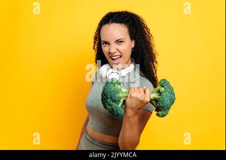 Happy excited active funny hispanic or brazilian curly woman in sport outfit, holds exercise with broccoli dumbbell while standing on isolated orange background, smiling, emotional facial expression Stock Photo