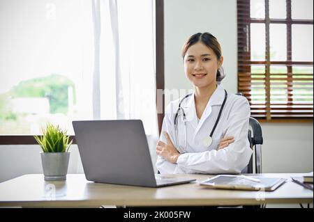 Attractive and confident young Asian female doctor sitting at her office desk, arms crossed and smiling to camera. Doctor, Specialist, Practitioner, p Stock Photo