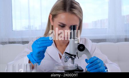 Female laboratory assistant analyzes samples using a microscope while working at a table in a laboratory, doing research Stock Photo