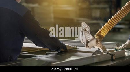 A male carpenter in the workshop saws chipboard and a wooden board with a circular saw on the machine. Furniture production. Stock Photo