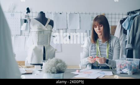 Young female tailor is busy choosing color of new garment drawn on sketch. She is picking different bobbins from box and applying them to drawing. Selecting sewing items concept. Stock Photo