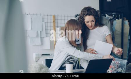 Team of two creative female designers is choosing color of sewing threads for garment drawn on sketch. Women are reeling bobbins in and out, talking and gesturing. Stock Photo