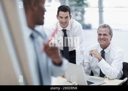 Brainstorming ideas. Businessmen smile and watch as a colleague writes on a flipchart. Stock Photo