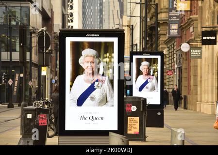Manchester, UK, 9th September, 2022.  People walk past electronic billboards featuring Her Majesty, Queen Elizabeth II, in Manchester, United Kingdom. Her Majesty, The Queen, died, aged 96, on 8th September, 2022. Manchester City Council has said on its website that the city of Manchester will be observing the official 10-day mourning period and that: 'Residents may wish to lay flowers to mark Her Majesty’s death. You can lay flowers at St Ann's Square'. Credit: Terry Waller/Alamy Live News Stock Photo