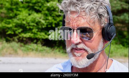 Gray-haired elderly man freelancer with a beard in sunglasses, working in the headphones on nature. A crazy and extraordinary old senior in the summer Stock Photo