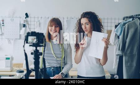 Young attractive vloggers clothing designers are recording video for their vlog. Women are showing garment sketches, gesturing and talking emotionally in tailoring studio. Stock Photo