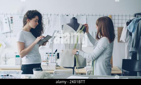 Professional seamstress is measuring patterns on mannequin with measure-tape while her coworker is working with tablet to write down measurement data. Light modern workshop in background. Stock Photo