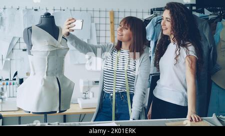 Two creative female designers are making selfie with smart phone while standing beside tailoring dummy in modern workplace. Attractive women are smiling and posing. Stock Photo