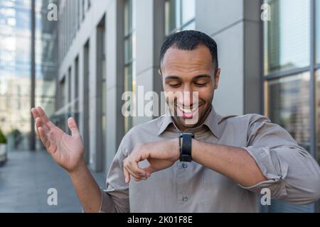 Young successful arab businessman talking on smart watch and smiling, app developer programmer walking outside modern office building, worker in casual shirt. Stock Photo