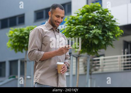 African American businessman smiling and happy walking outside the office building, holding a cup of hot drink during a break, and using the phone, browsing web pages and typing messages Stock Photo