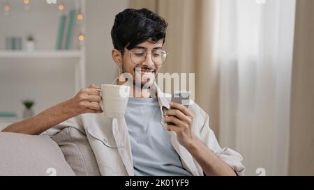 Millennial arabic guy indian handsome 30s man sitting at home on couch drinking hot tea coffee cappuccino cocoa from cup receive message looking at Stock Photo
