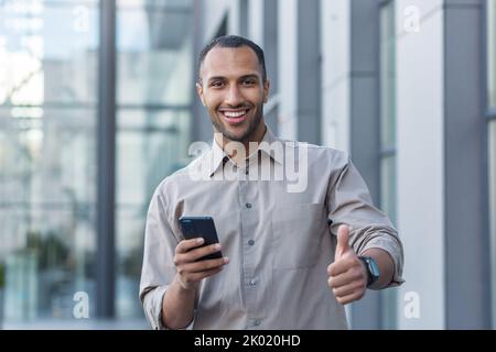 Young African American businessman outside office building smiling and looking at camera, thumbs up, programmer using smartphone for app testing. Stock Photo