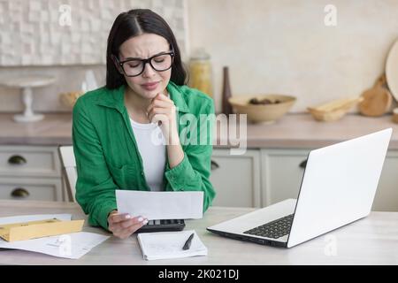 Divorce. A young beautiful woman received divorce papers. Shocked, upset. Sitting in the kitchen at home, wearing glasses and a green shirt, holding a letter in his hands. Stock Photo