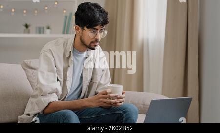 Young freelancer male business man bearded guy in glasses drinks tea coffee delicious drinking at home sitting on couch watching movie in laptop Stock Photo