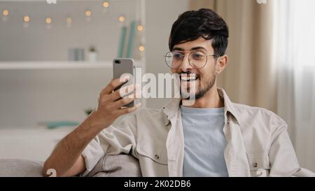 Portrait indoor male arabic guy unshaven man with glasses sitting on couch at home chatting online using mobile phone reads news scrolling browsing Stock Photo