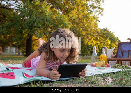 A little girl uses a tablet while on a picnic in a park, children and education with technology Stock Photo