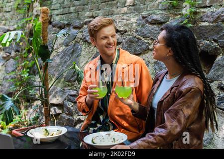 cheerful multiethnic friends holding glasses with cocktail while having lunch near laptop in cafe on terrace,stock image Stock Photo