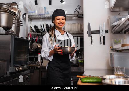 cheerful and young african american chef holding bowl with salad in professional kitchen,stock image Stock Photo