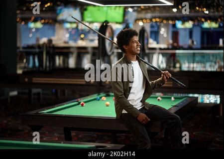 Young pool player with cue sitting on billiard table Stock Photo