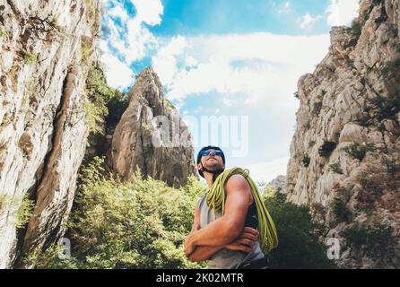 Smiling climber man in protective helmet and sunglasses with climbing rope on the shoulder in the Paklenica between rock cliff walls. Active extreme s Stock Photo