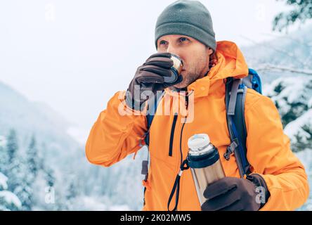 Man drinking a hot drink from thermos flask dressed bright orange softshell jacket while he trekking winter mountains route. Active people in the natu Stock Photo