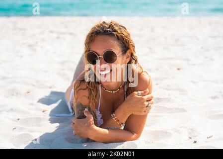 Portrait of cheerful young adult woman laying at the beach on white sand. Tropical summer holiday vacation. Relaxed female people smile and enjoy the sun. Blue sea water in background. Travel leisure Stock Photo