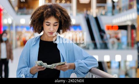 Millennial african american girl shopper consumer client woman standing in shopping business center counting money cash dollars banknotes earning Stock Photo