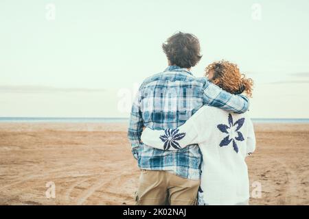 Rear view portrait of couple in love admiring the beach and ocean hugging with love. Romantic life in winter holiday vacation. People in relationship embracing. Love and togetherness concept Stock Photo
