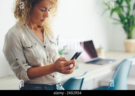 Pretty young adult woman sending message on mobile phone. Concept of social connection with smartphone. Female people using technology at office. Lady work at home. Modern workplace in background. Stock Photo