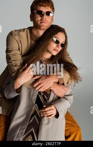 redhead young man in stylish sunglasses hugging girlfriend isolated on grey,stock image Stock Photo