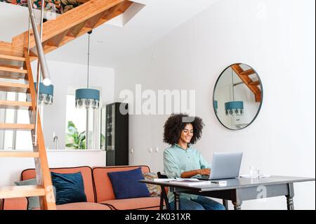 Side view of black woman sitting with laptop computer at the table at cozy living room. Young woman typing on keyboard, working online Stock Photo