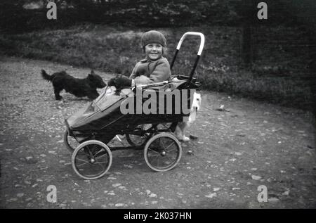 1932, historical, outside on a stoney path, a young girl wearing a coat and beret and with a lovely smile on her face, sitting in a metal bodied pushchair of the era, with the family's scottie dogs near her on the path, England, UK Stock Photo