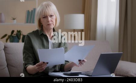 Caucasian old sad homeowner housewife makes woman budget checks monthly income by checking paper notes using app laptop. Middle-aged businesswoman Stock Photo