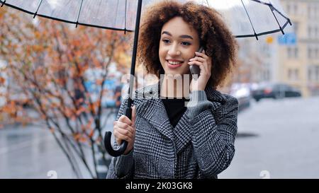 Happy active friendly curly haired girl african american woman with transparent umbrella in city in windy rainy weather answering call talking on Stock Photo
