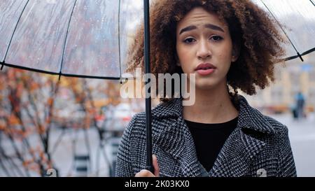 African American girl curly-haired sad woman in stylish coat stands in autumn on city street with transparent umbrella in rain suffers from cold winds Stock Photo