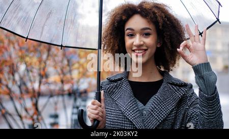 Beautiful happy successful lady curly african american woman girl with transparent umbrella stands in autumn city in rain looking at camera smiling Stock Photo