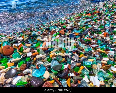 Natural polish textured sea glass and stones on the seashore. Azure clear sea water with waves. Green, blue shiny glass with multi-colored sea pebbles Stock Photo