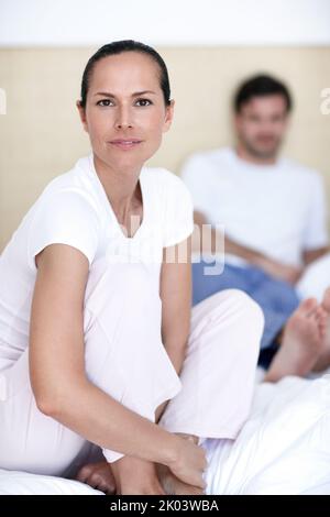 Ready to start her morning routine. A young woman sitting cross-legged on a bed with her husband in the background. Stock Photo