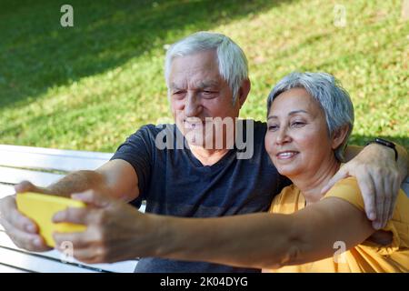 Happy senior spouses take selfie picture sit on bench in summer park. Grey-haired couple look at cell phone screen make videocall outdoors use modern Stock Photo