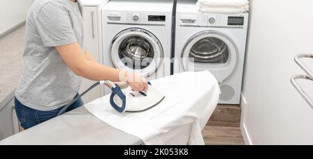 Woman ironing white shirt on board in laundry room with washing machine on background Stock Photo