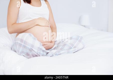 Holding her future. Cropped image of a young woman holding her pregnant belly on her bed. Stock Photo