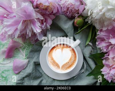 Flat lay cup of coffee with heart shape pattern surrounded with light pink peony flower petals on green textile background Stock Photo