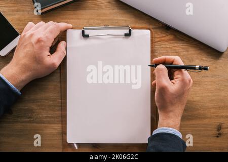 Business mockup, layout. Top view of clipboard with white sheet of paper vertically placed on wooden table. Male hands are holding pencil or ballpoint Stock Photo