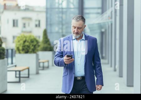 Gray-haired senior handsome man businessman in suit walking near office center on street, holding mobile phone, typing message, checking mail. Stock Photo