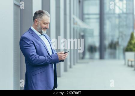 Senior handsome businessman man stands on the street near the office center and waits for customers, employees, holds the phone in his hands, dials a number, messages. Stock Photo